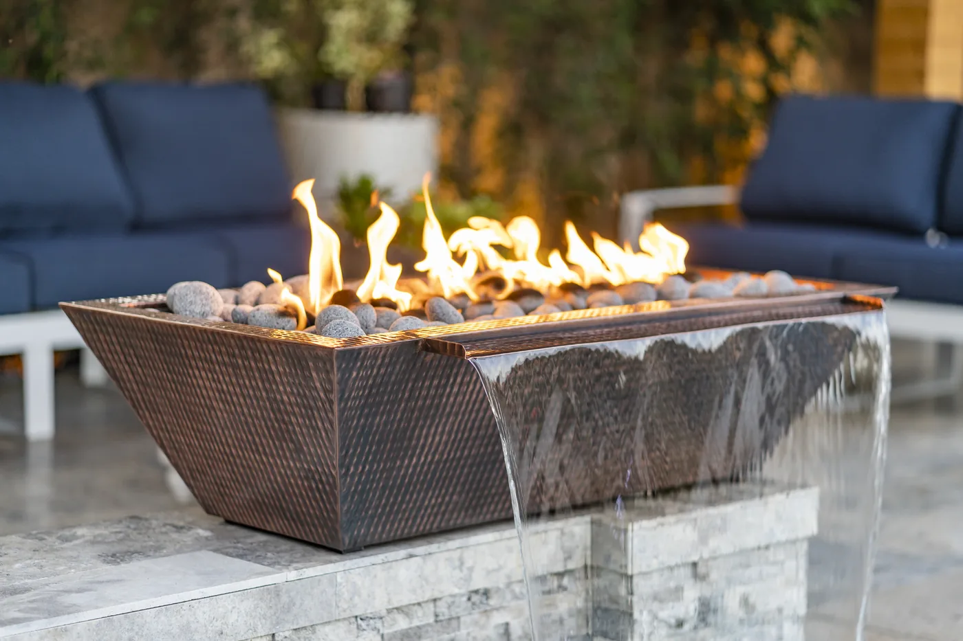 A hammered copper linear fire and water bowl, flame burning across the top while a sheet of water spills from the lip