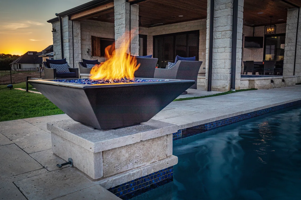 A square GFRC chestnut fire bowl burning on a travertine pedestal at the edge of a pool, with a covered patio behind it at sunset